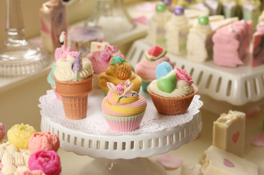 variety of dessert shaped soap sitting on a cake plate with other cast soaps surrounding it like flowers, fruit, macaroons, and ice cream in a variety of colors